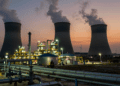 Natural gas power plant at dusk with steam cooling towers, industrial pipelines, and a utility worker on the horizon